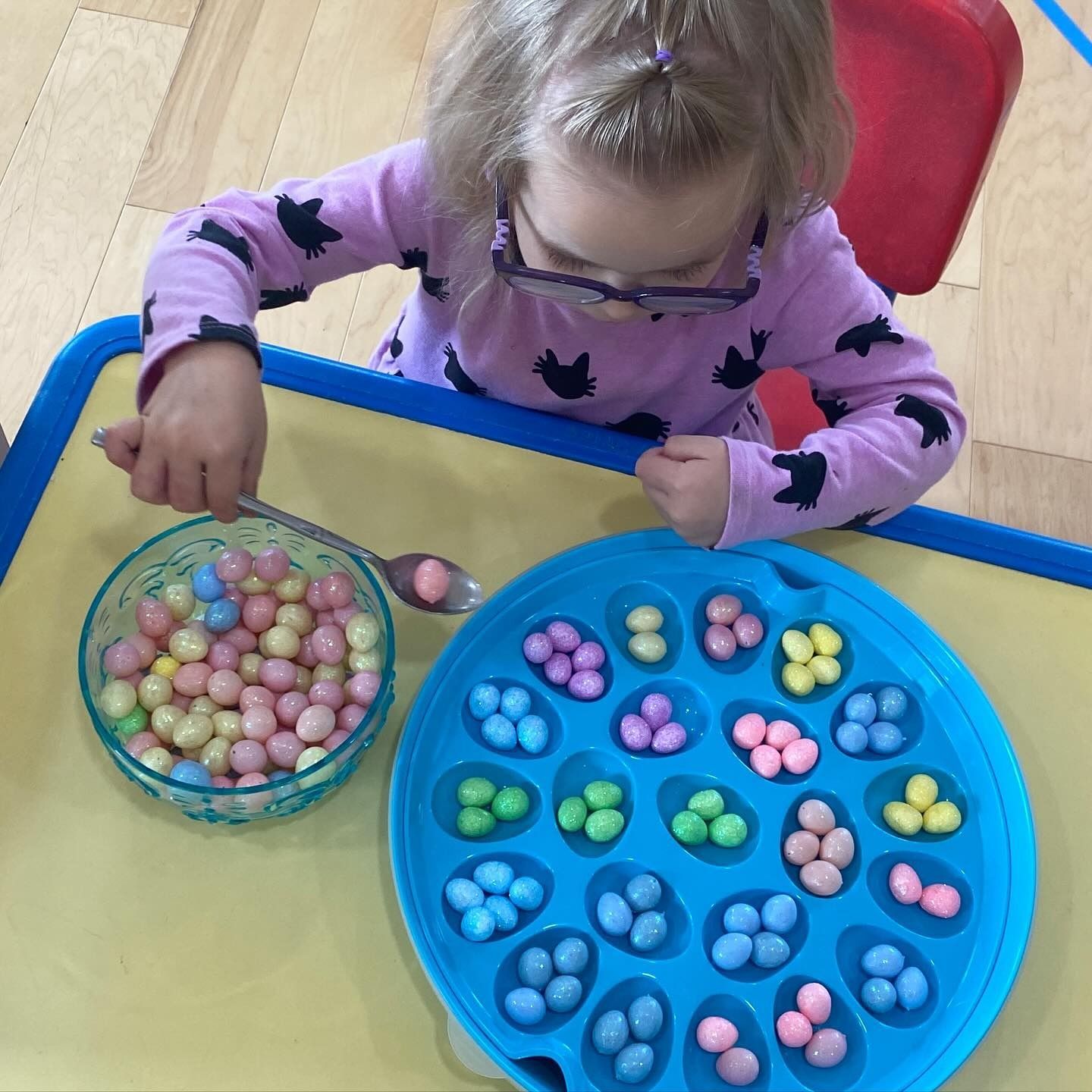 Child with glasses sorting colorful candies into a blue tray with a spoon.