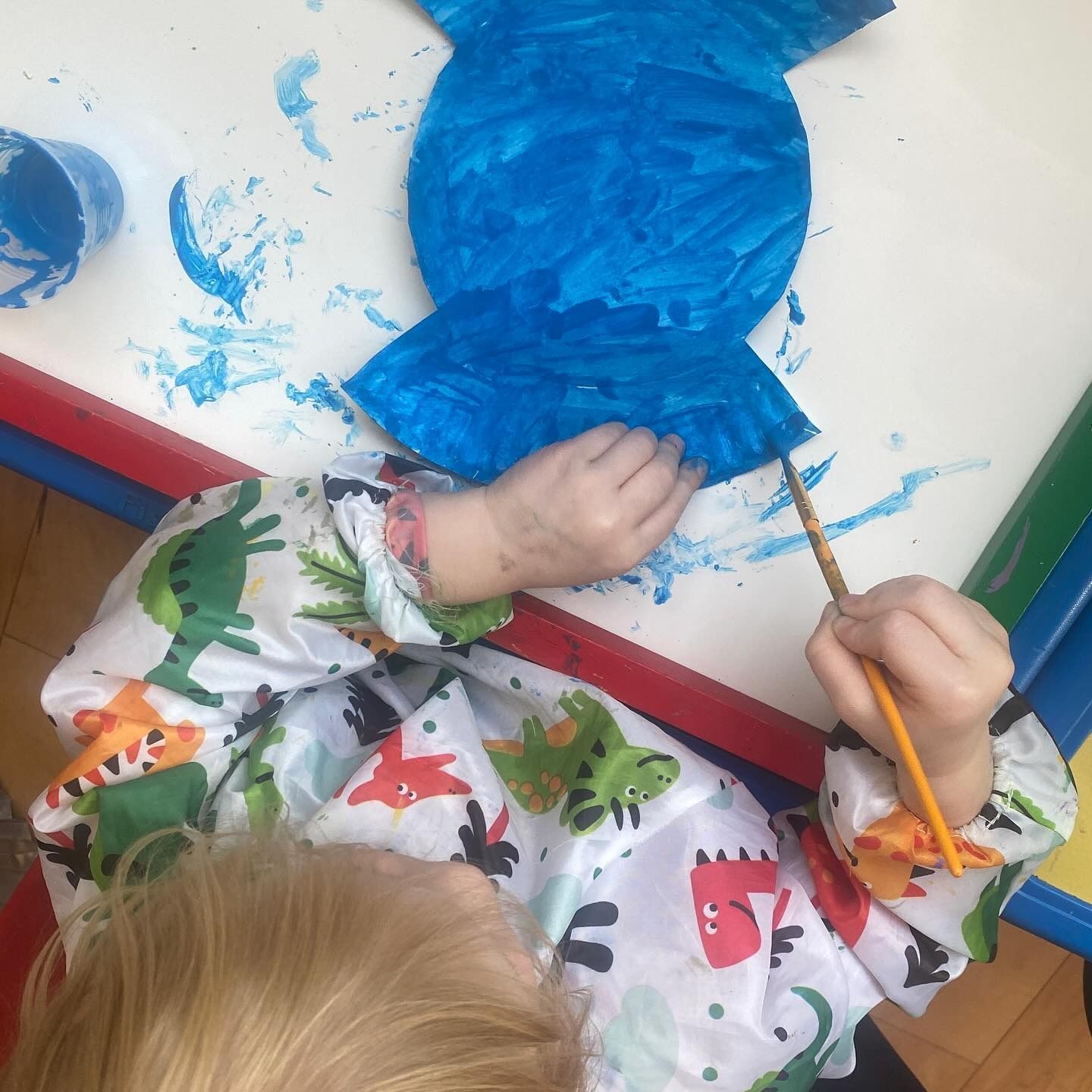 Child in dinosaur art smock paints a blue paper cutout at a table, using a paintbrush.