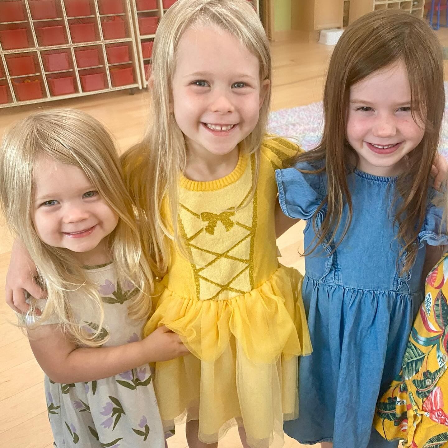 Three smiling girls, one in a yellow dress, pose indoors.