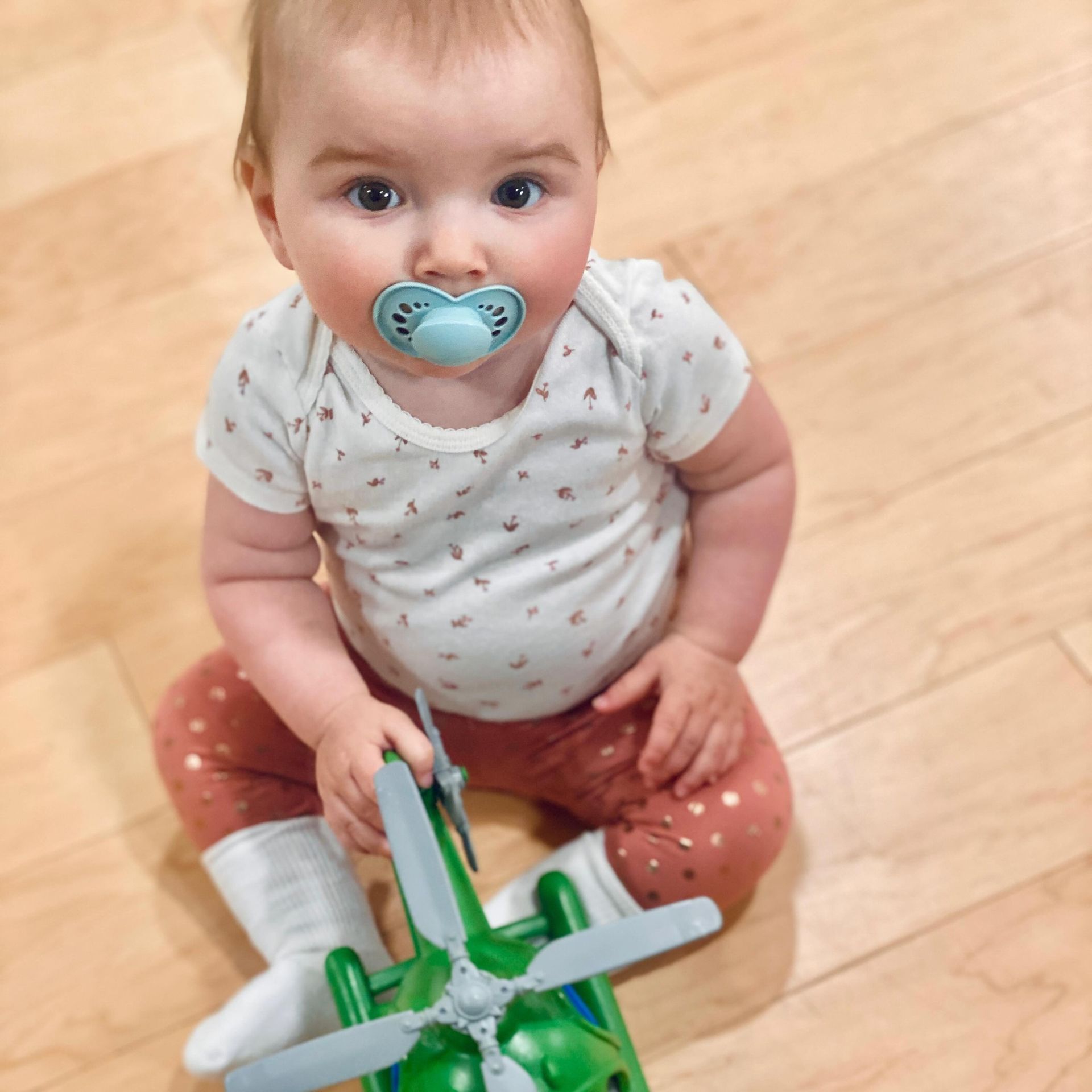 Baby with pacifier holds a green toy helicopter, sitting on a wooden floor.
