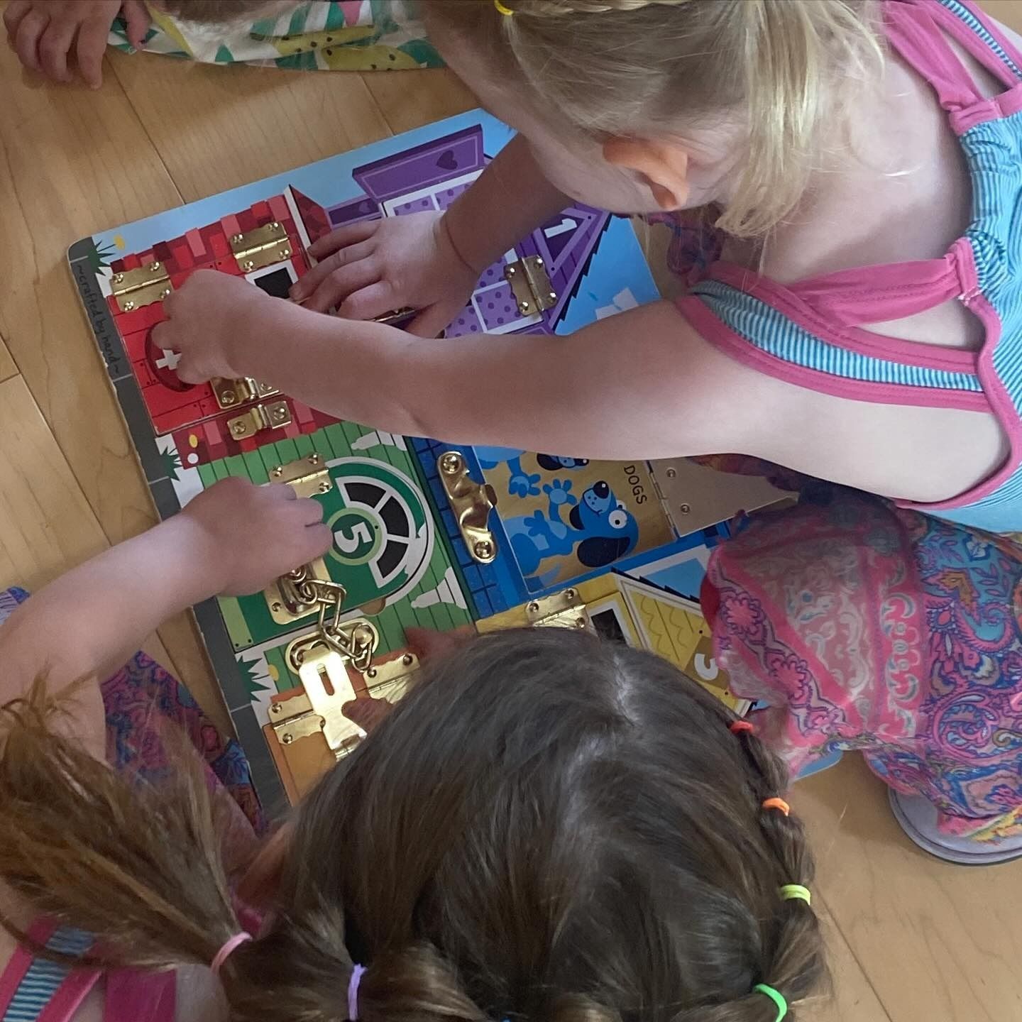 Two children playing with a multi-lock toy on a wooden floor, reaching and manipulating locks.