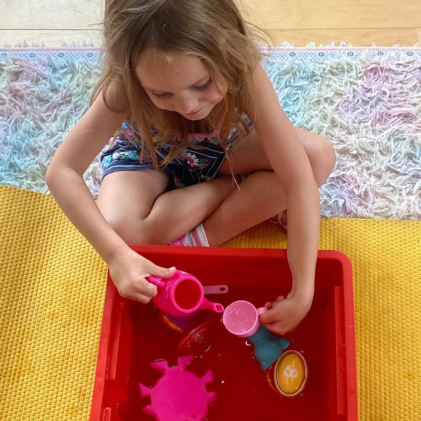 Young child playing with toys in red bin, sitting on colorful rug.