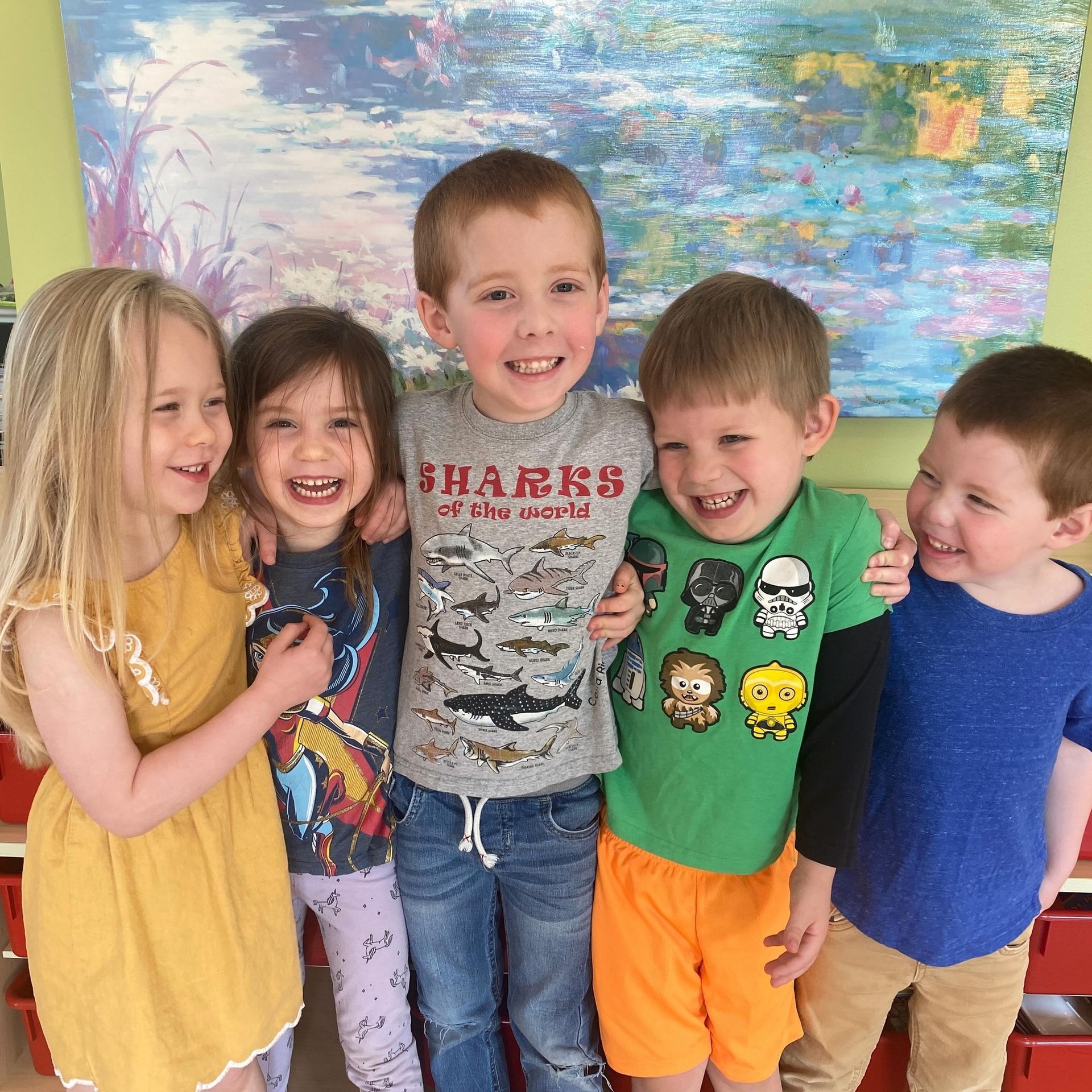 Five children smiling, arm-in-arm. They stand in front of a colorful abstract painting.