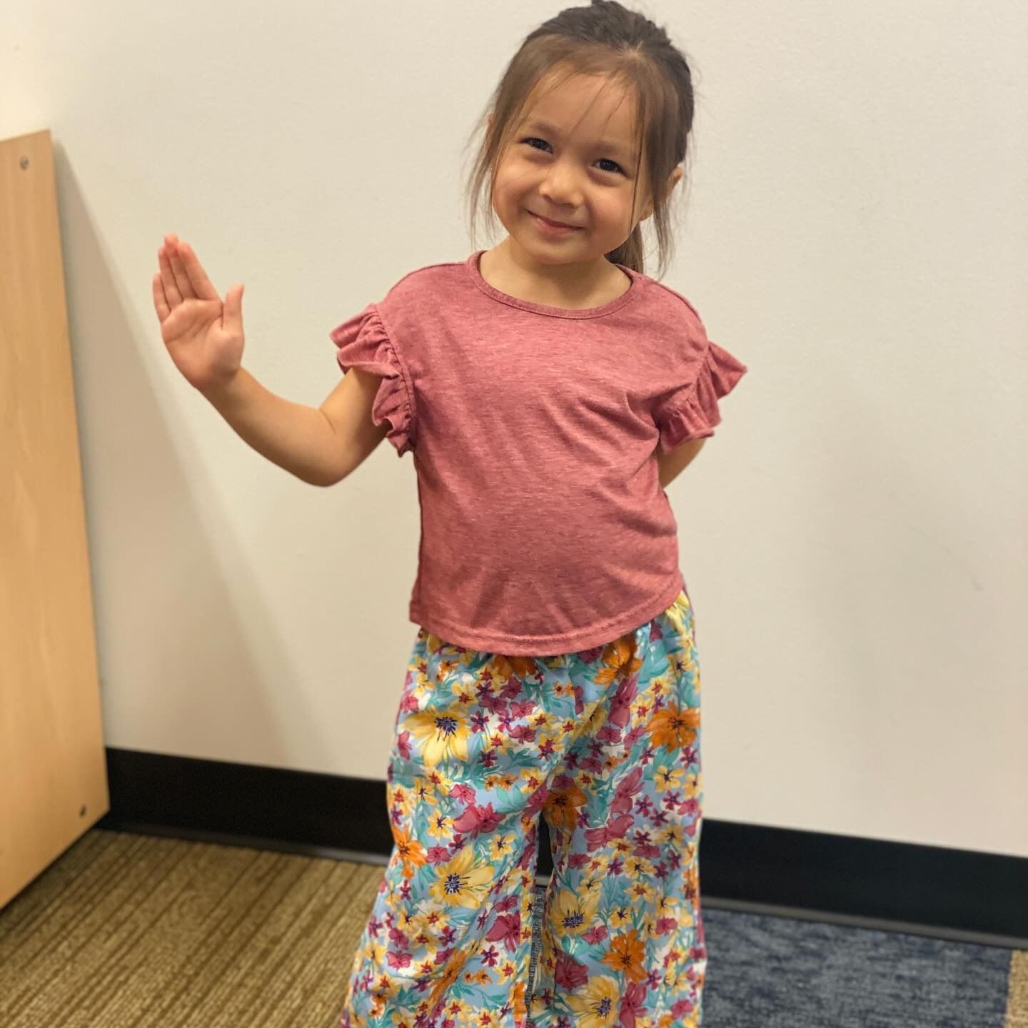 Young girl in floral pants and red shirt waving in a room.