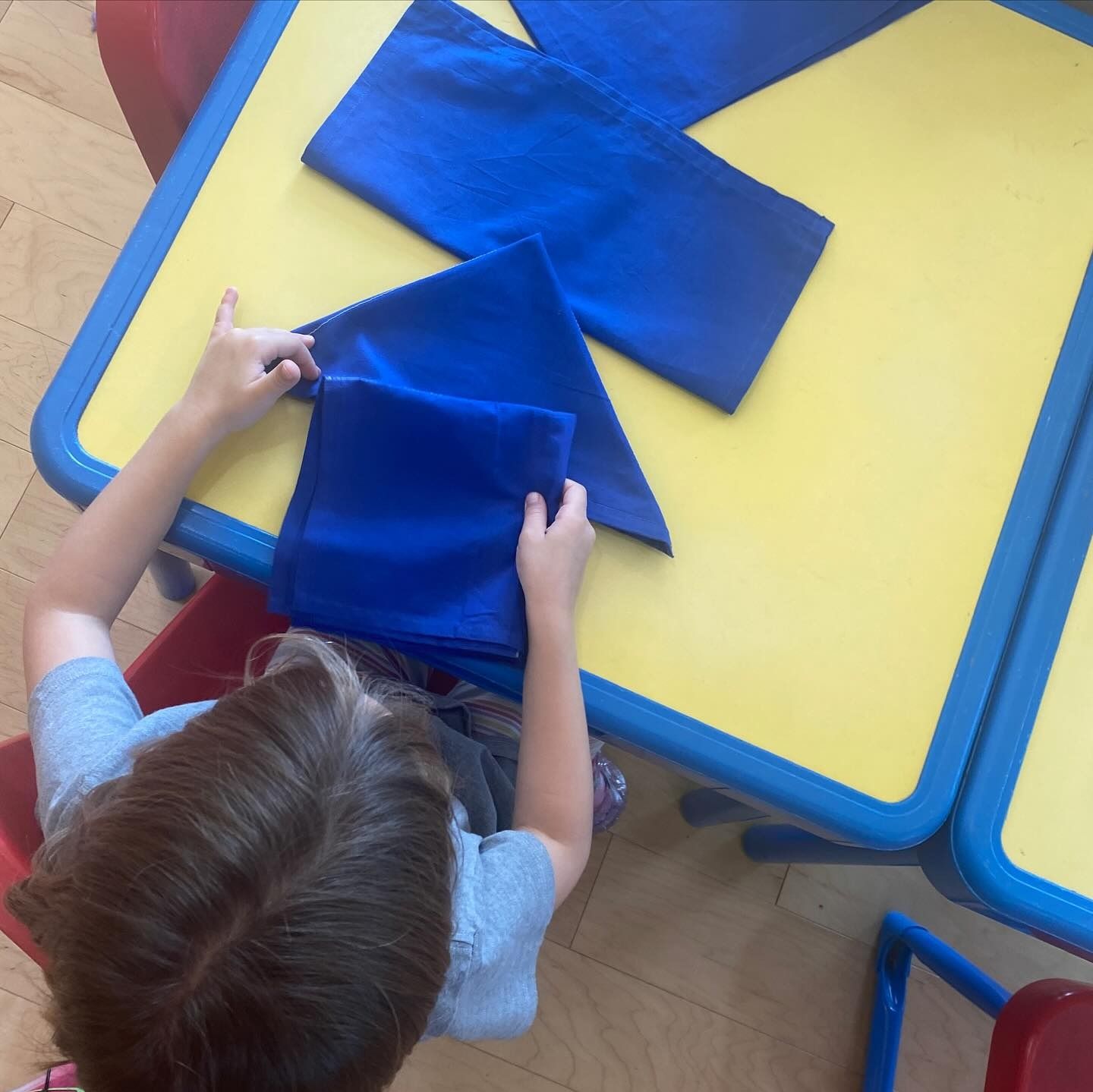 Child folding blue fabric on a yellow table.