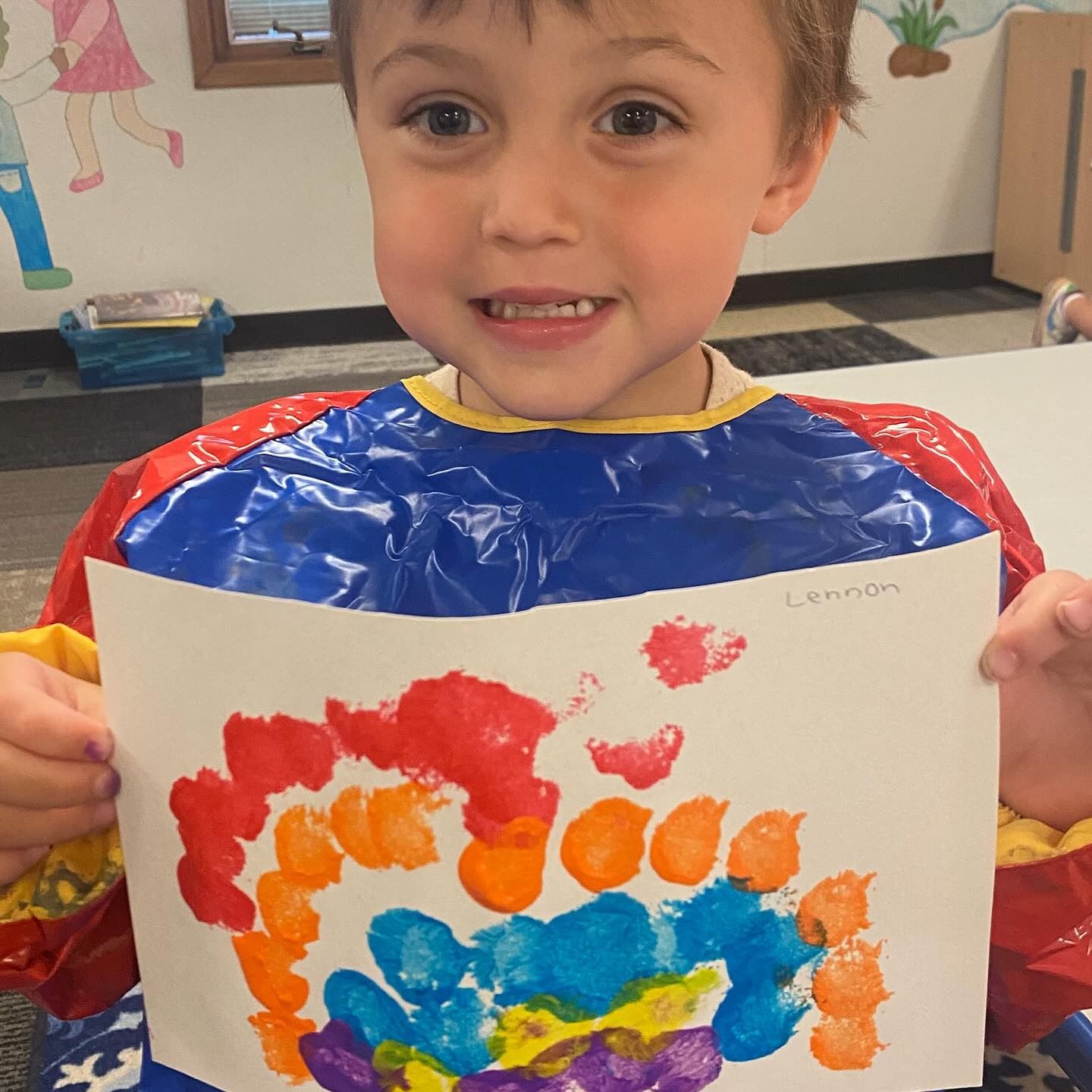 Boy holding a colorful thumbprint rainbow art, smiling in a classroom setting.