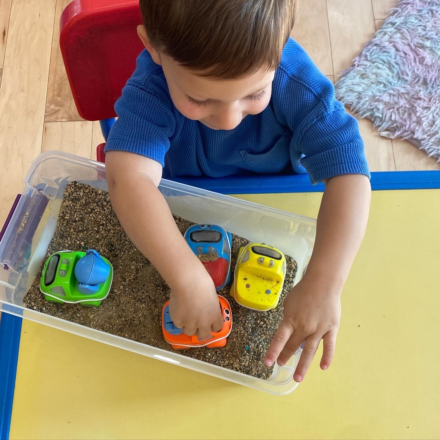 Child playing with toy cars in a sand tray; yellow table, blue shirt, wood floor.