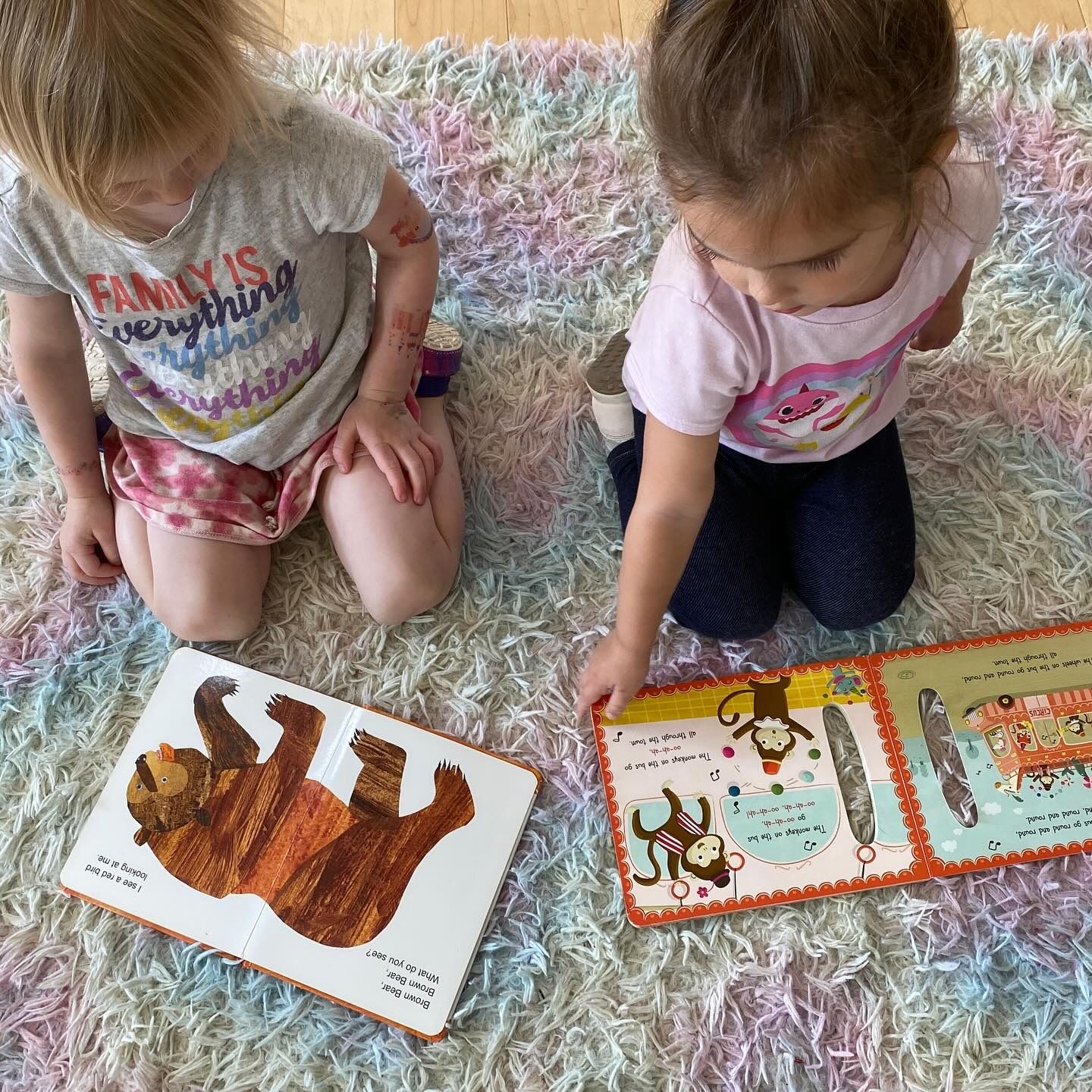 Two children kneeling on a fluffy rug, looking at open books with illustrations.