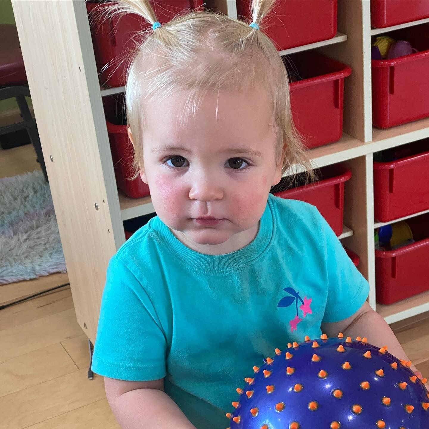 Blonde toddler with pigtails holding a blue spiky ball, in a room with red bins.