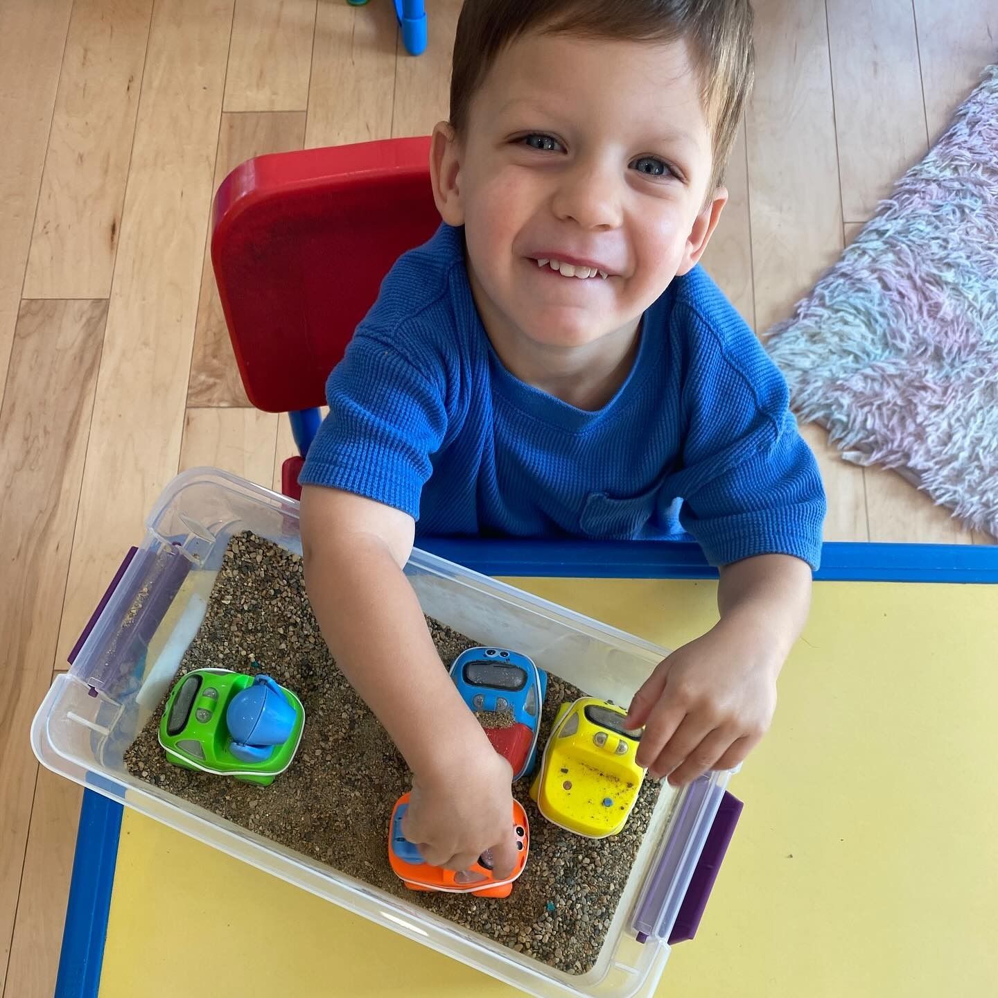 Boy playing with toy cars in a sensory bin, smiling at table.