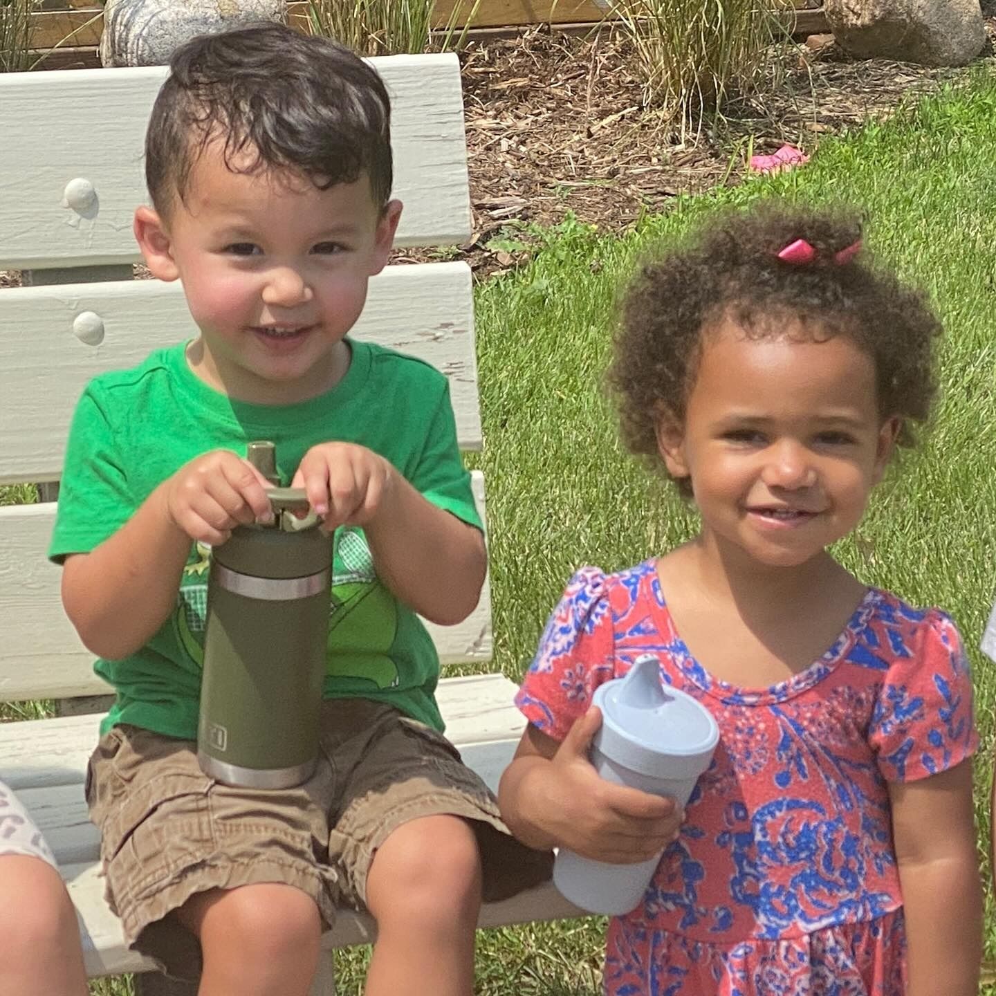 Two children sitting on a bench, smiling, and holding water bottles. Green shirt and pink dress.
