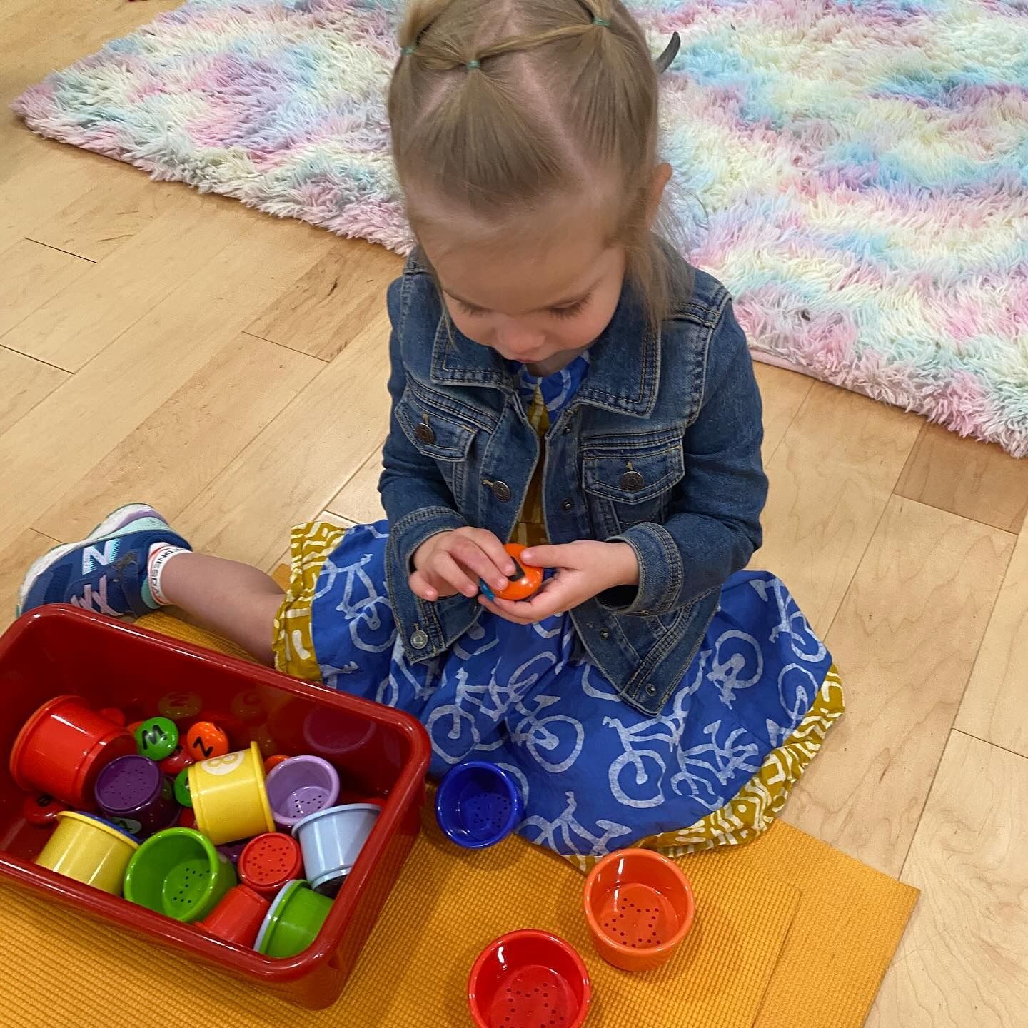 Young child sitting on floor with colorful cups and objects.