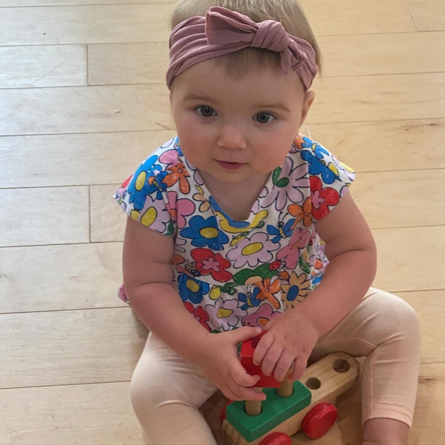 Baby wearing a floral top and pink headband, playing with a wooden toy train.