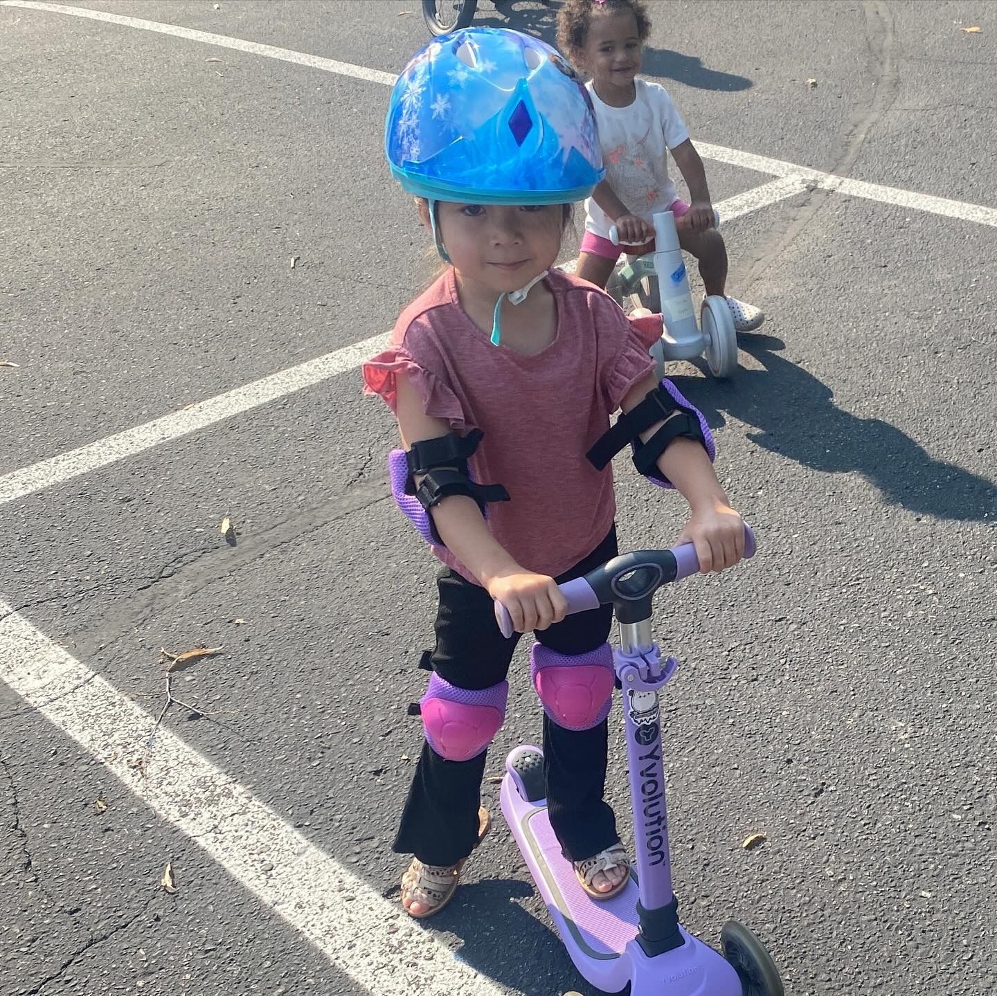 Girl on a purple scooter, wearing a helmet and pads, smiles. Another child rides a scooter in the background.