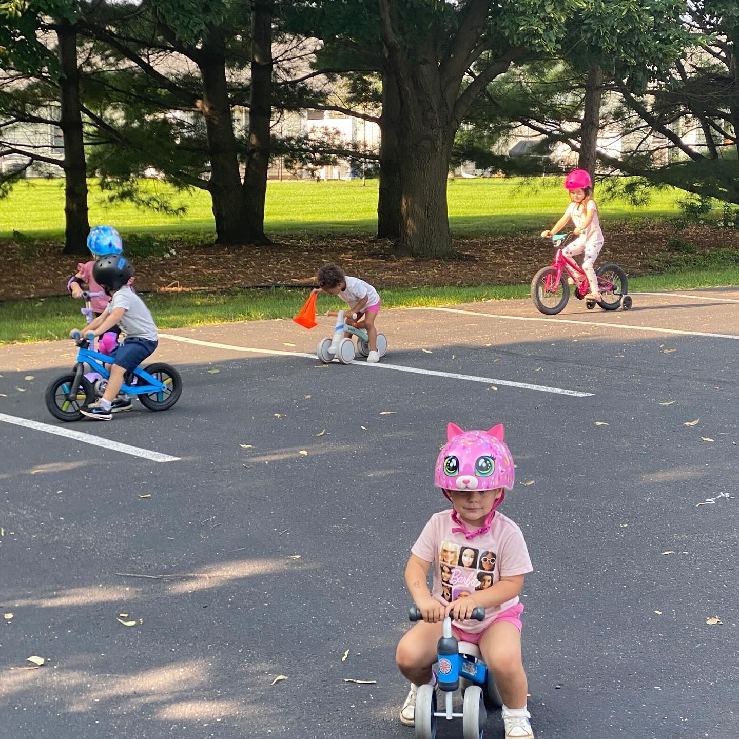 Children riding balance bikes and a tricycle on asphalt, wearing helmets.