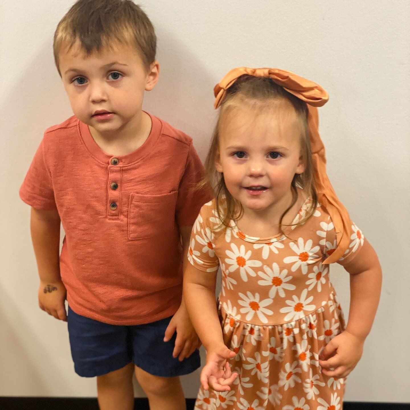 Boy in orange shirt and shorts stands next to a girl in a floral dress with a bow on her head.