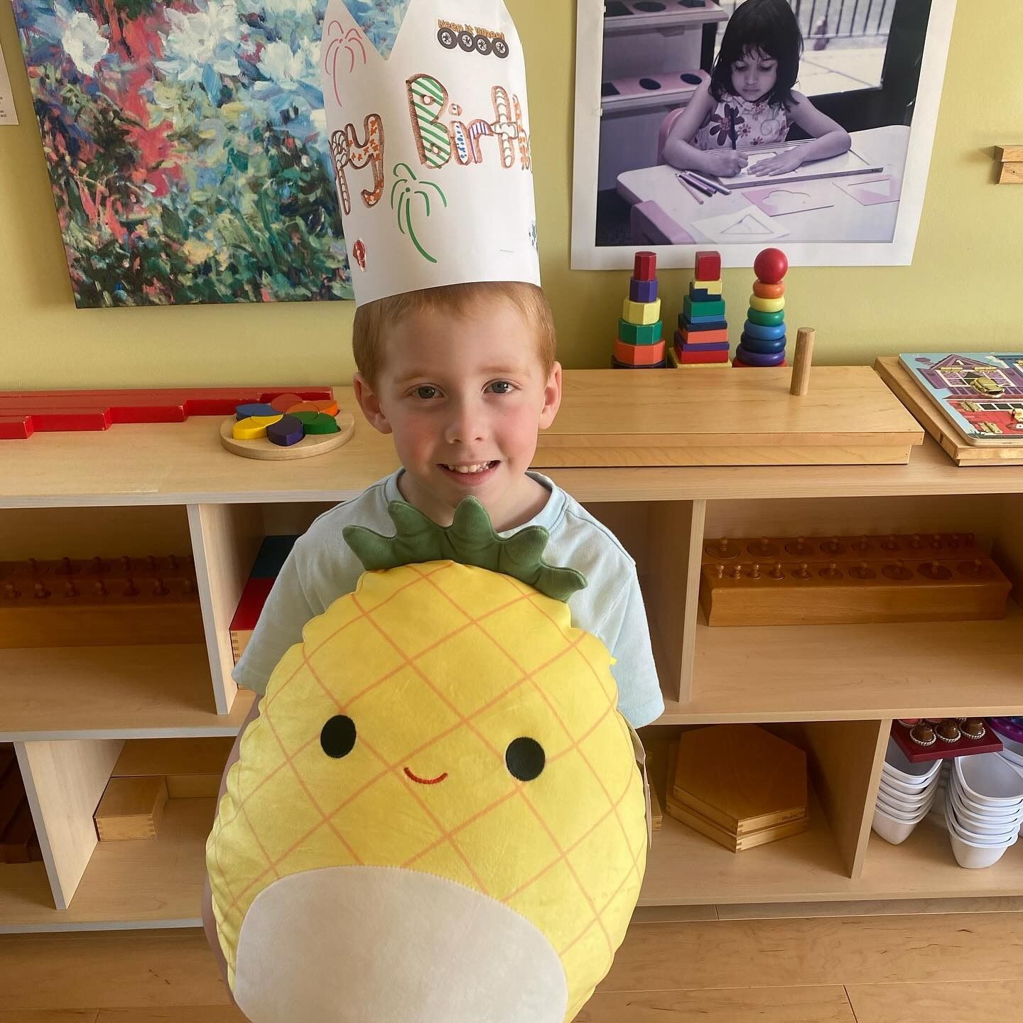Boy wearing a chef hat and holding a pineapple plushie, smiling in a classroom setting.