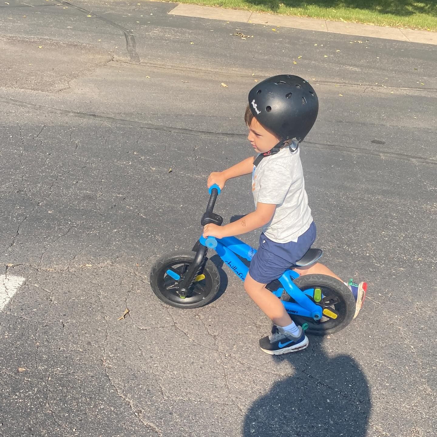 Child in blue shorts and white shirt on a blue balance bike wearing a black helmet on pavement.