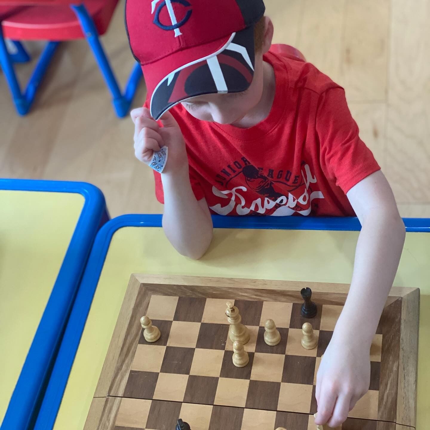 Boy in red shirt and Twins hat playing chess at a yellow table.