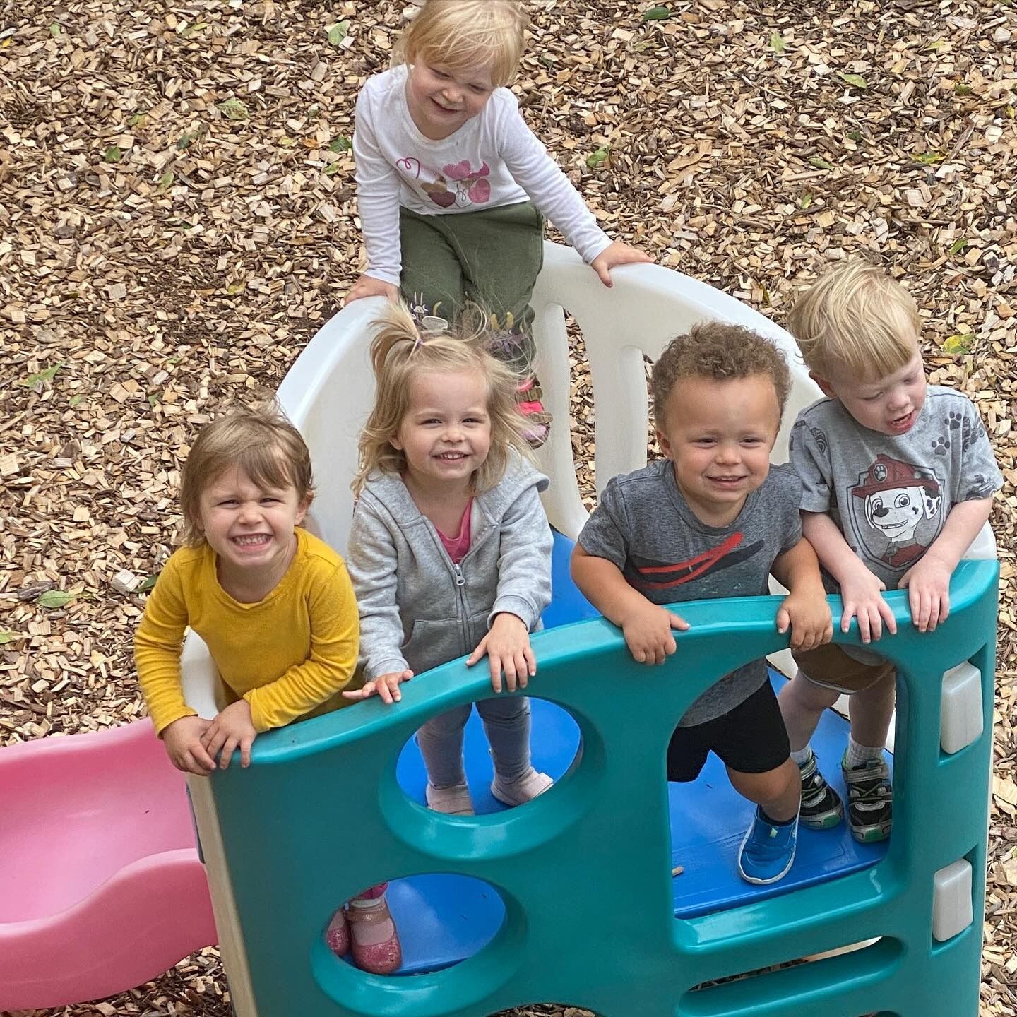 Children playing on a colorful play structure; smiling and looking at the camera. Outdoor setting.