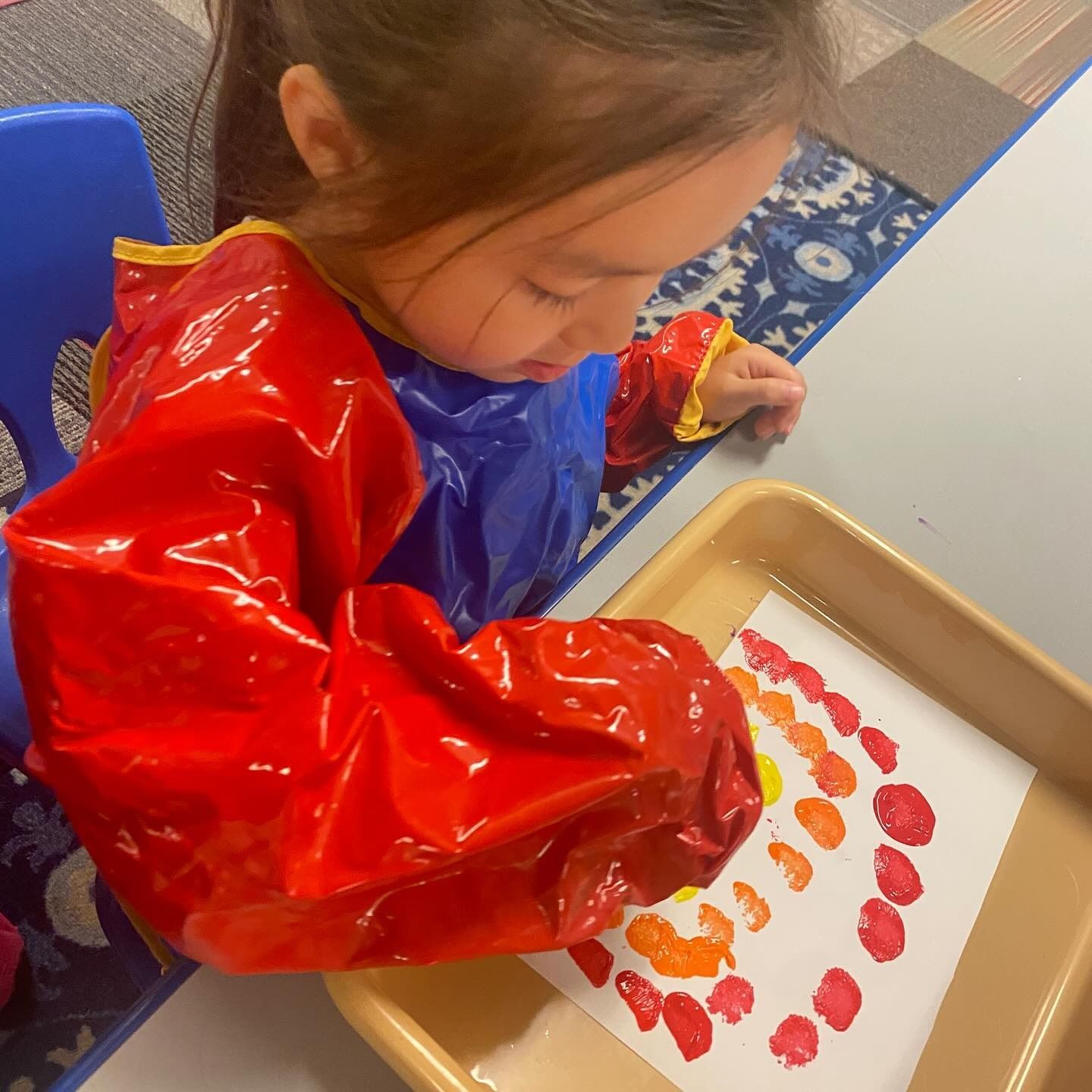 Child wearing art smock painting dots on paper in a tray.