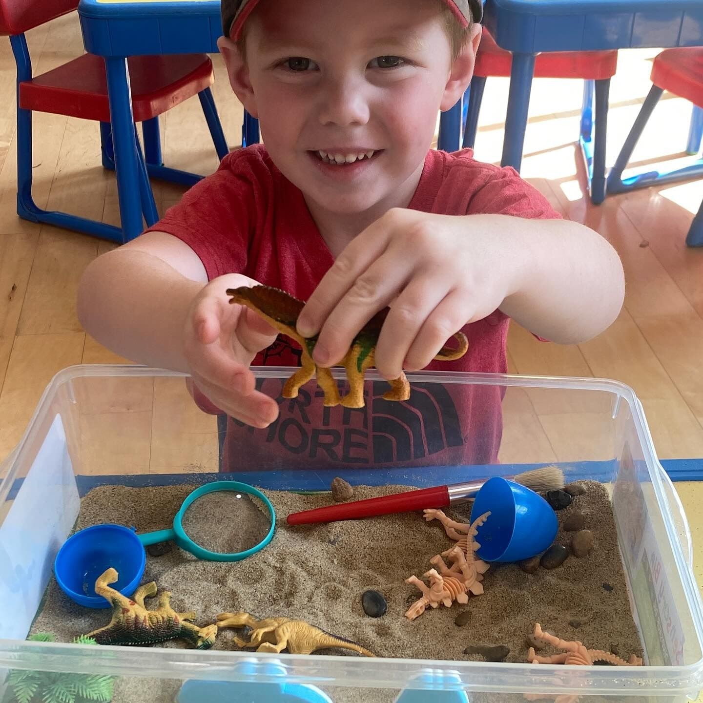 Boy smiles, holding dinosaur toy over a sandbox. Blue scoop, magnifying glass, and more toy dinos are visible.