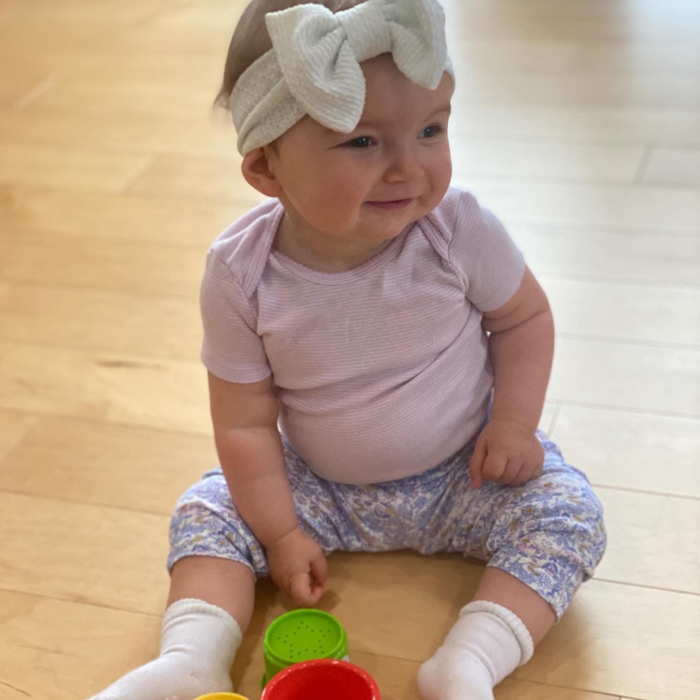 Smiling baby wearing a bow, pink onesie, and floral pants sits with colorful toys on a wooden floor.