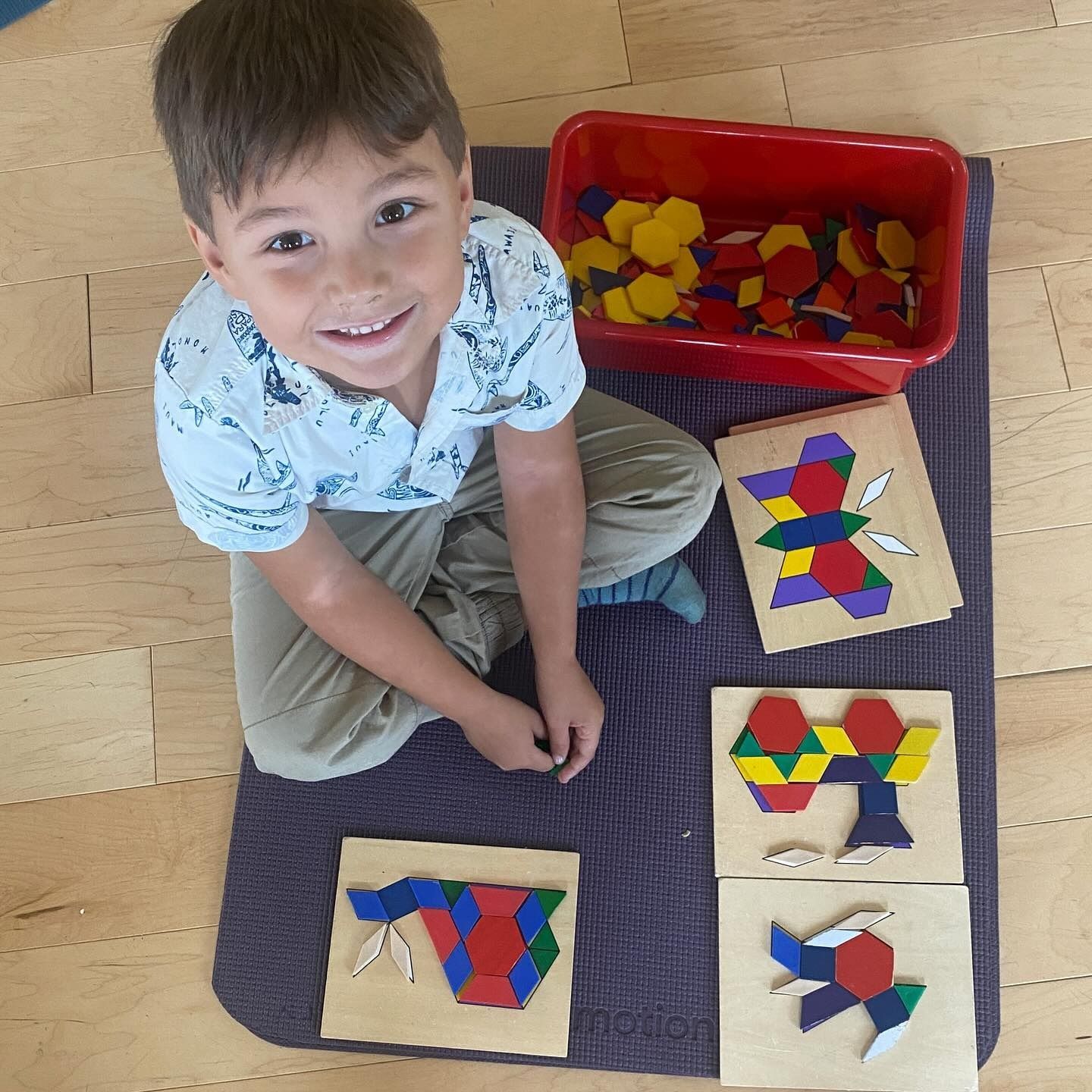 Boy smiling, working with colorful geometric shapes.  Red bin of shapes,  wooden puzzles.  Sitting on mat.