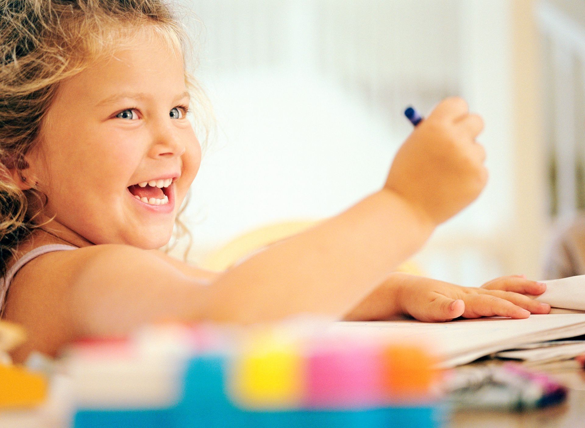 Smiling child with curly hair drawing with a crayon at a table.