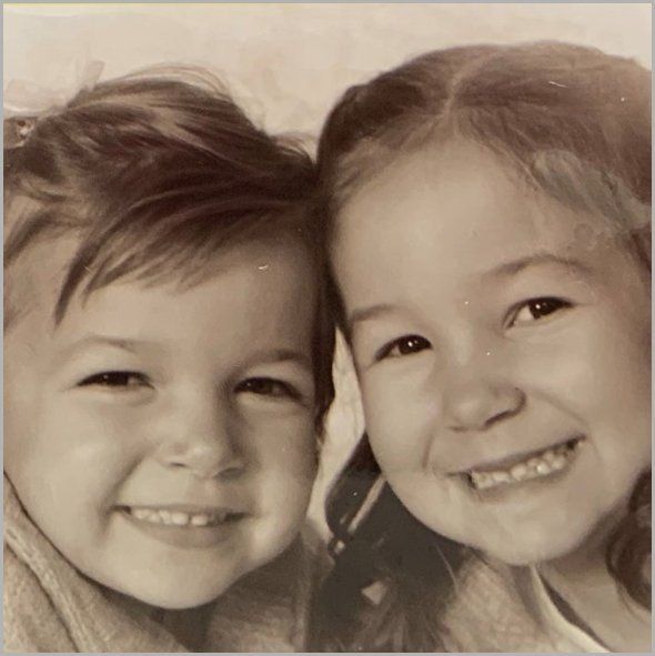 Two children smiling, close-up shot. One with side-swept hair, the other with a ponytail. Black and white.