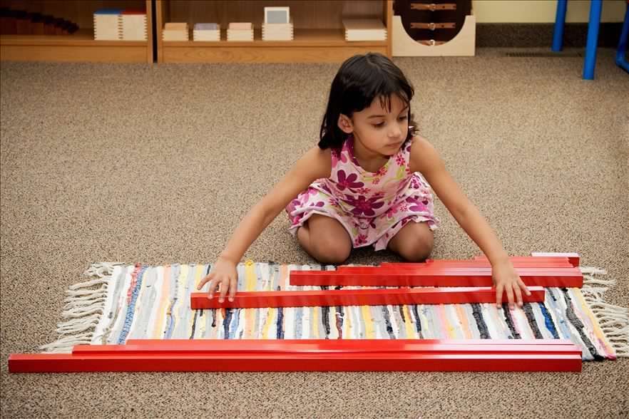 Girl arranging red wooden rods on a patterned mat in a Montessori classroom.