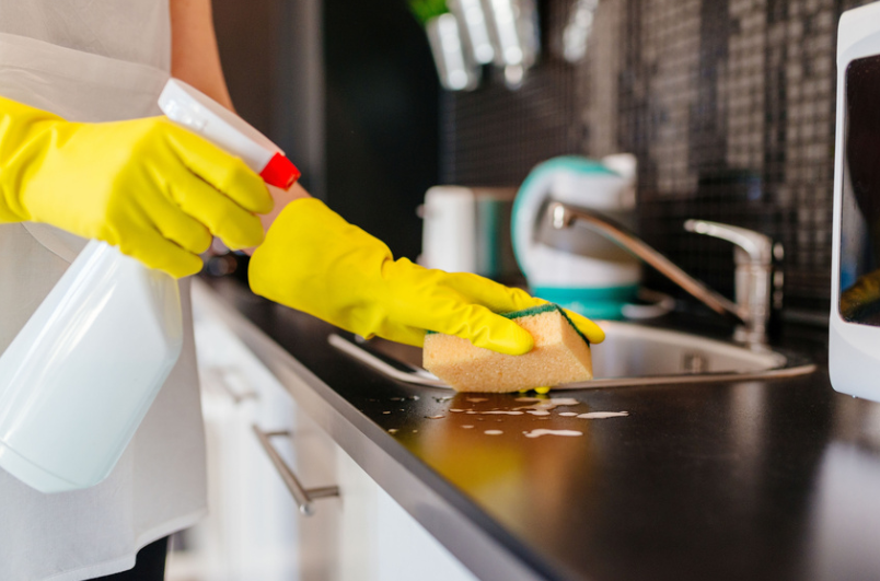 Person in yellow gloves cleaning a black countertop with a sponge and spray bottle in a kitchen.