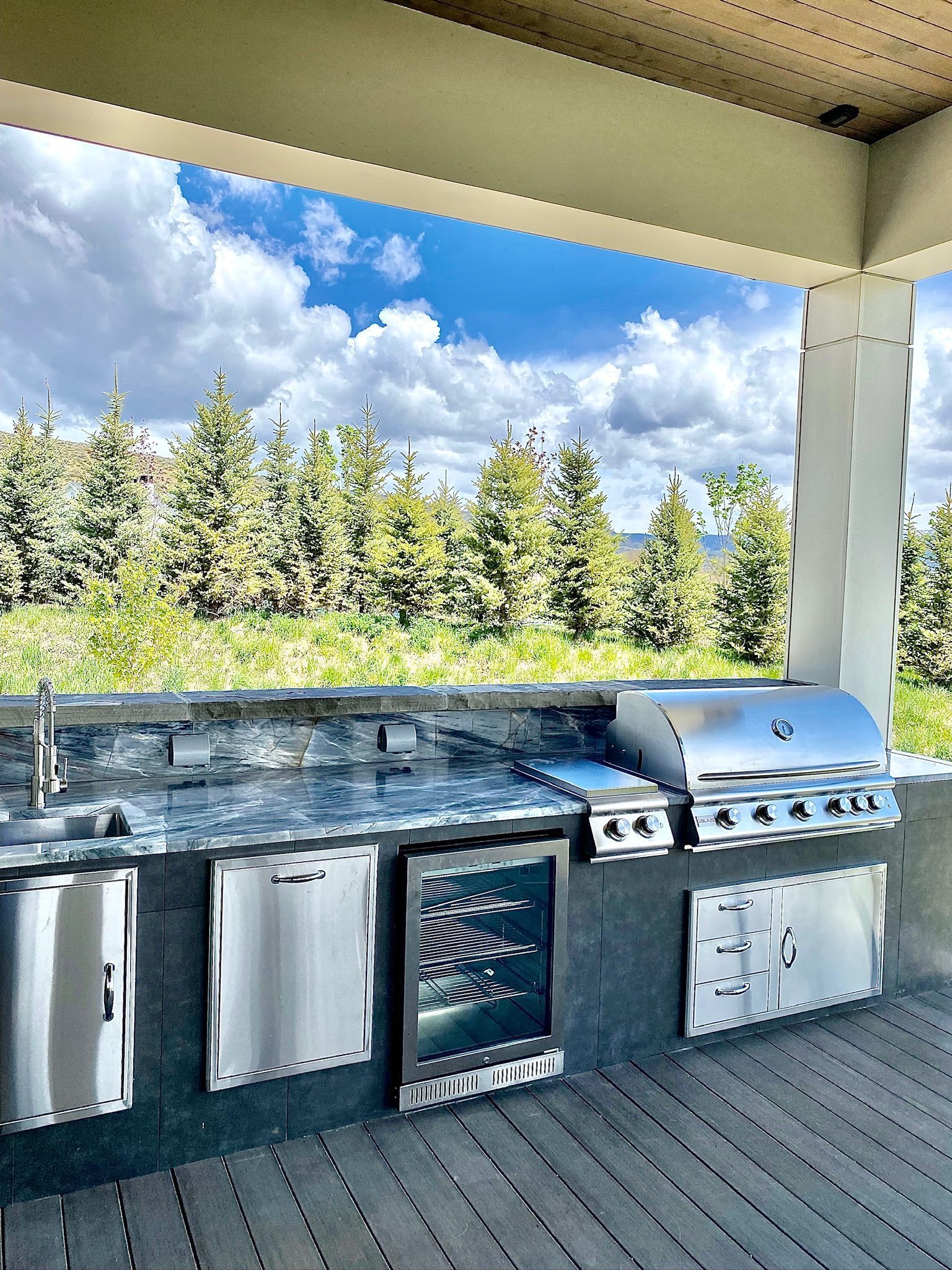 Outdoor kitchen with stainless steel grill, cabinets, and a refrigerator, overlooking a forest