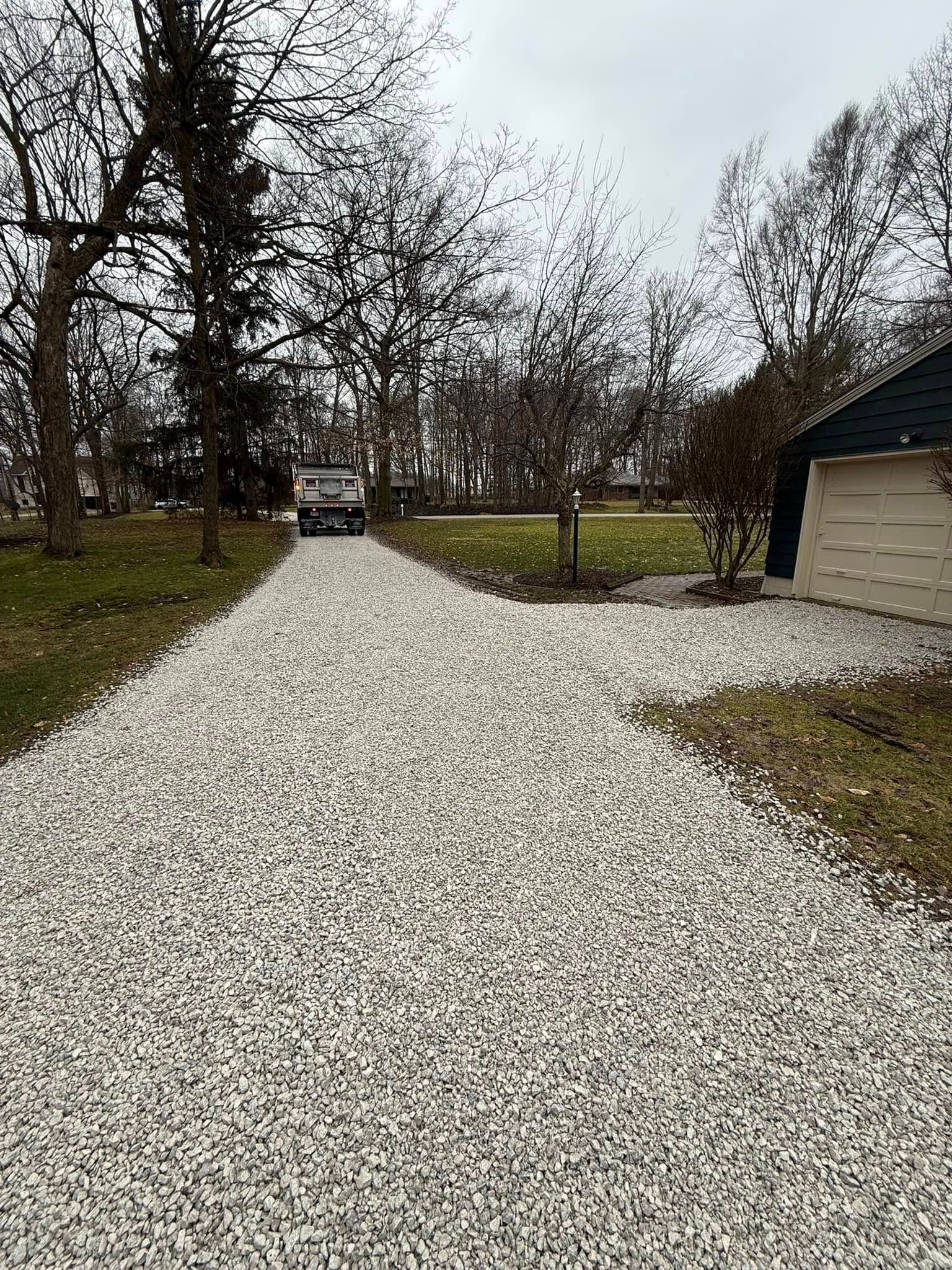 Gravel driveway lined with leafless trees leads to a parked car beside a beige building under an overcast sky