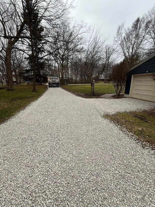 Gravel driveway lined with leafless trees leads to a parked car beside a beige building under an overcast sky