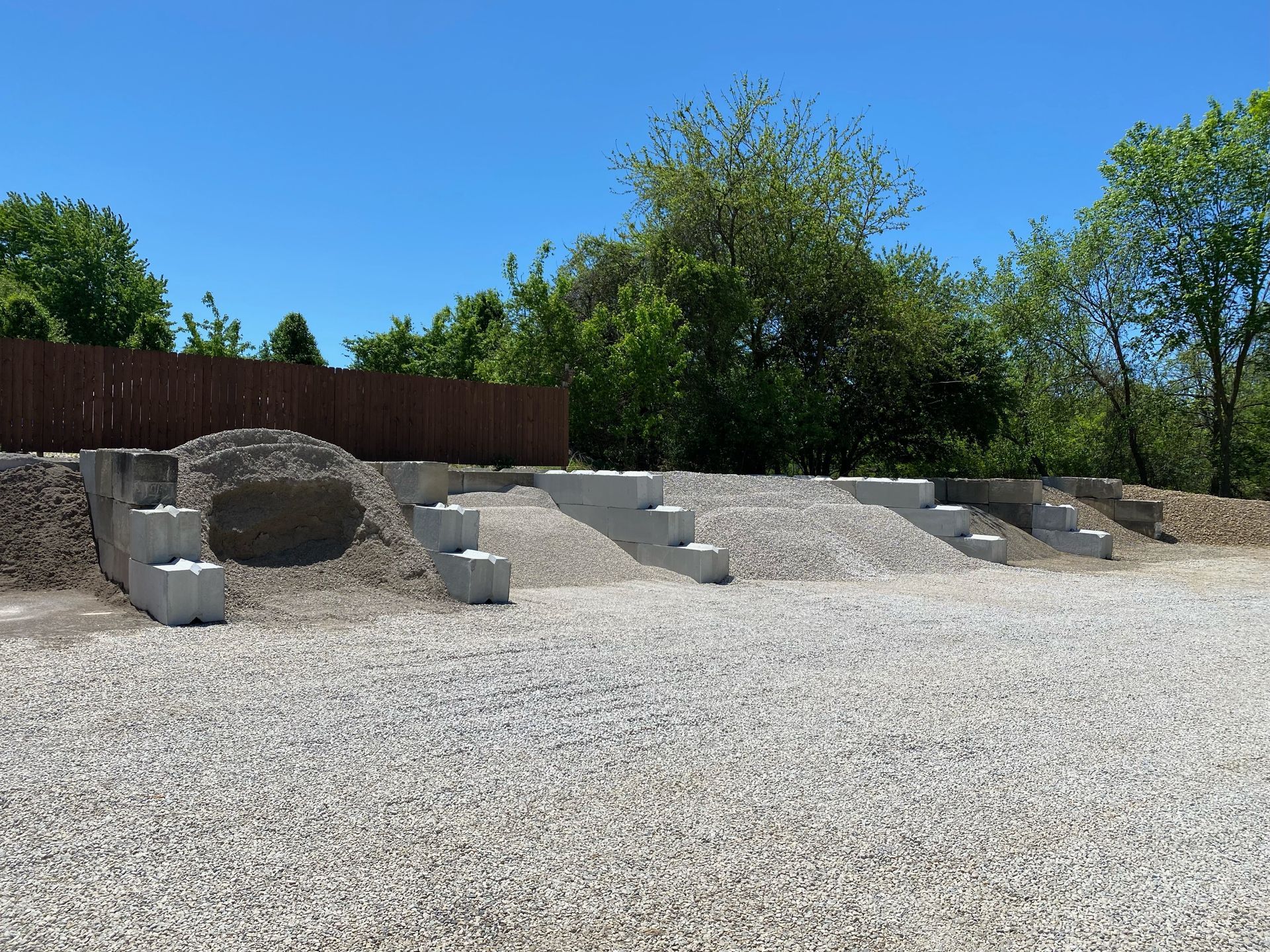 Gravel yard with a stone fire pit, stepped retaining wall, and trees under a blue sky