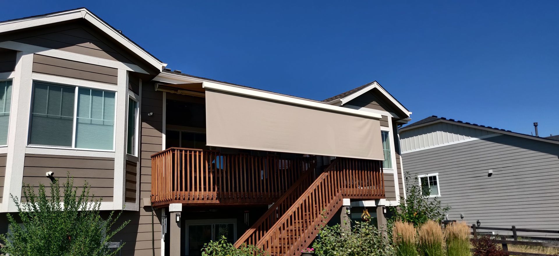 A two-story house with a wooden deck and a tan awning under a clear blue sky.
