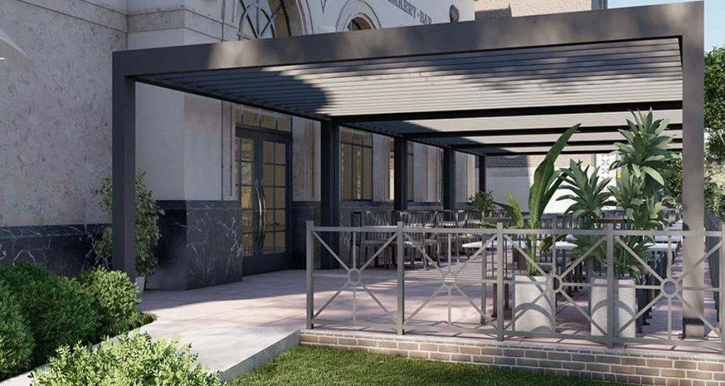 Pergola with outdoor seating next to a building. Gray frame, plants, tables.