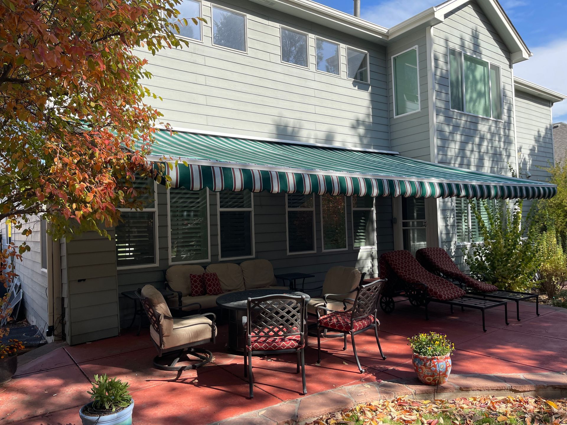 Back patio with green-striped awning, outdoor furniture, red concrete, and a two-story gray house.