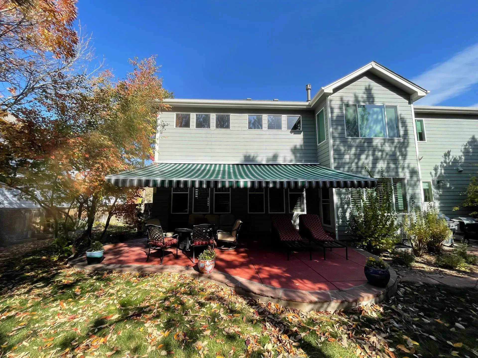 Back of a two-story gray house with a green and white striped awning over a patio. Autumn leaves and blue sky.