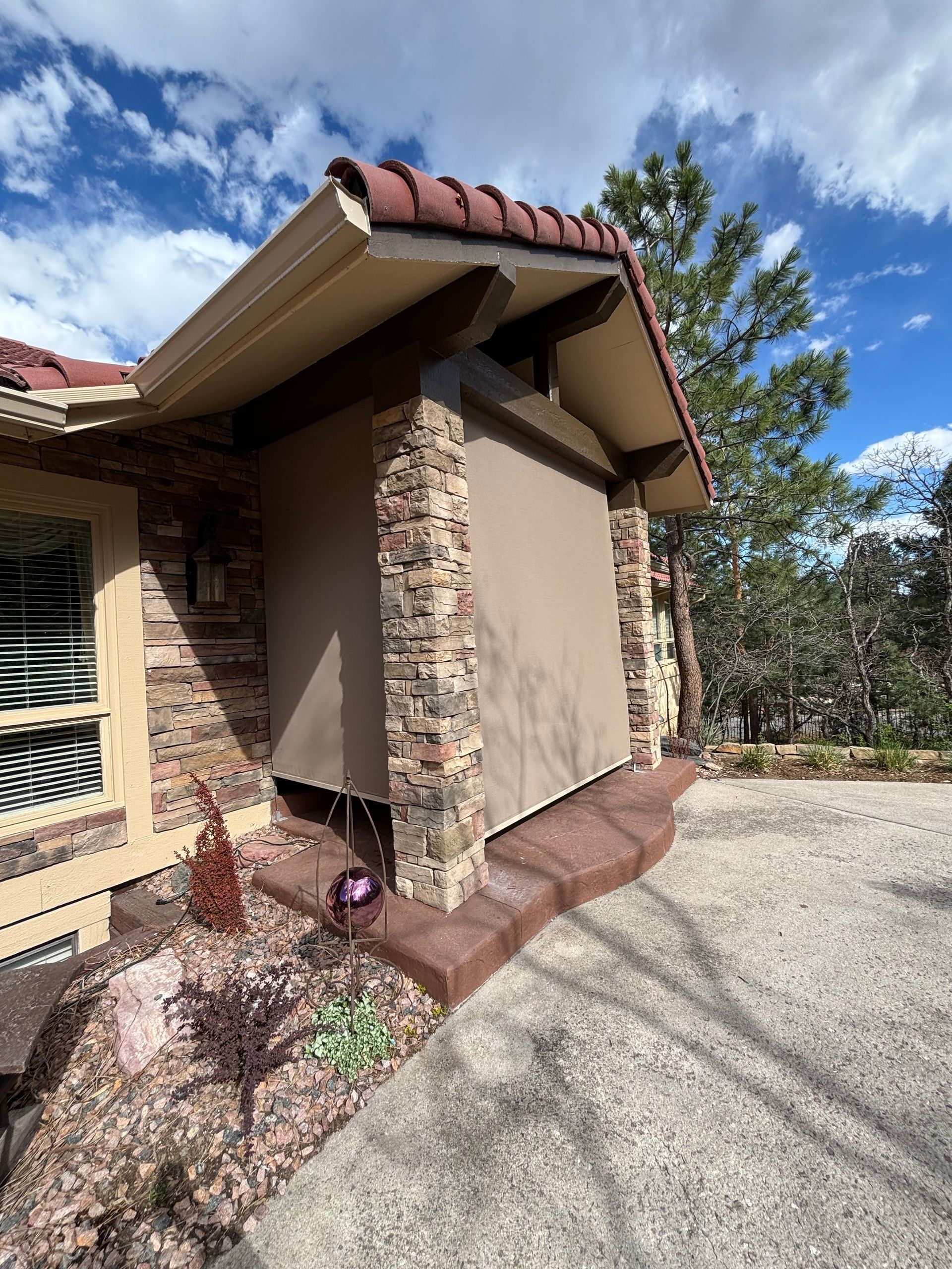 Exterior of a house with stone columns, red tile roof, and a retractable brown shade.