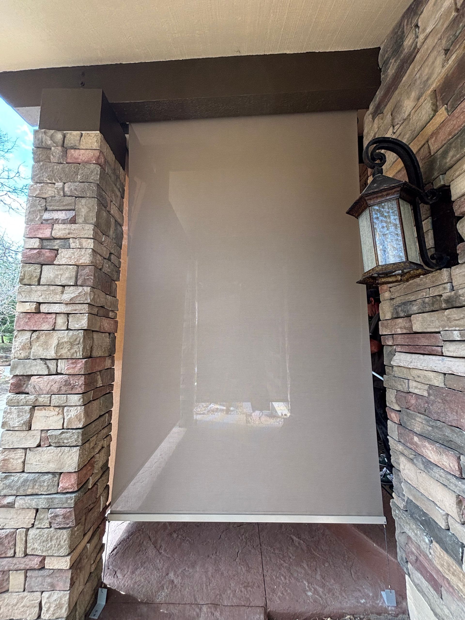 Beige sun shade rolled down on a porch, framed by stone columns and a lantern.