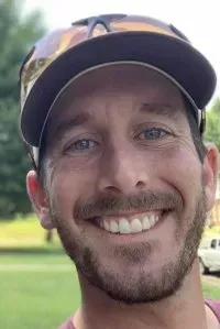 Man with a beard and baseball cap smiles broadly outdoors.
