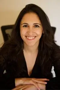 Woman with long dark hair smiles, sitting at a desk. She wears a dark top and appears to be indoors.