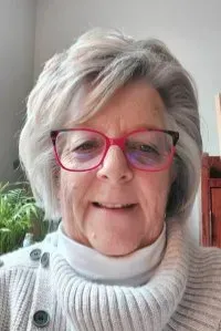 Woman with gray hair and red glasses smiles, wearing a white turtleneck and sweater.