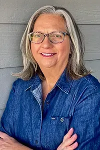 Woman with glasses, gray hair, and denim shirt smiling with arms crossed.