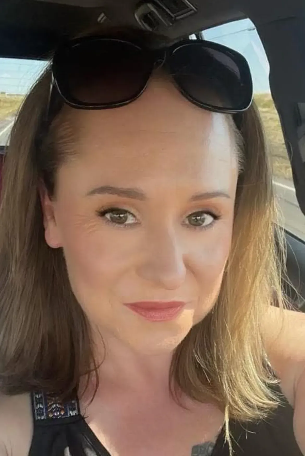 Woman wearing sunglasses, looking at the camera. Light skin, brown hair, inside a vehicle.