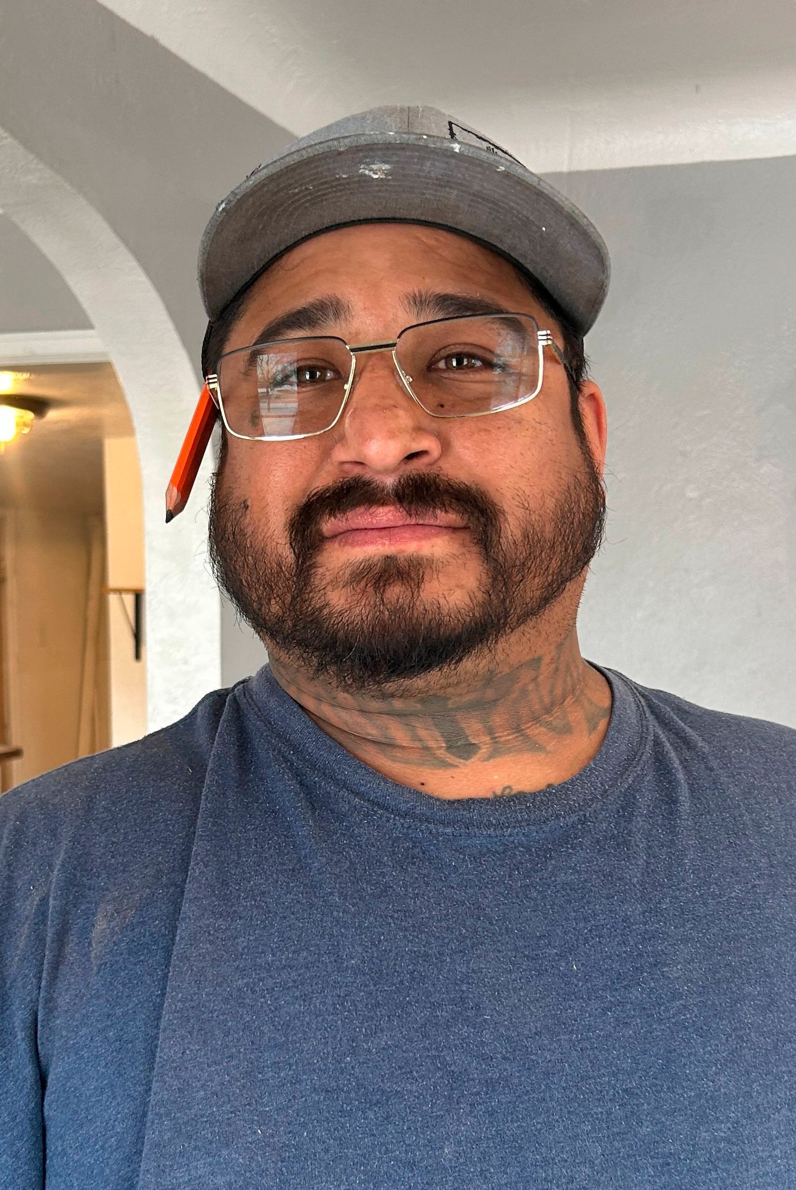 Man with glasses and a black hat smiling, wearing a black shirt, outside near a light-colored wall.