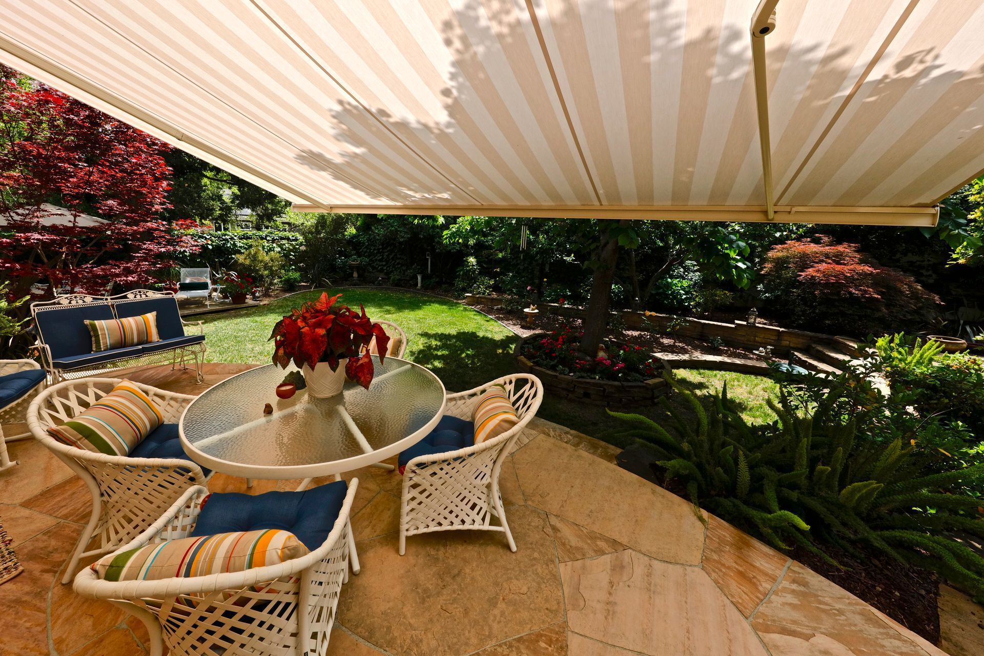 Patio with striped awning, glass table, wicker chairs with blue cushions, and a garden view.