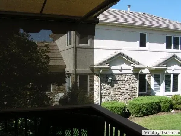 View of a two-story house through a dark shade. The house has light stucco and stone facade. Sunny day.